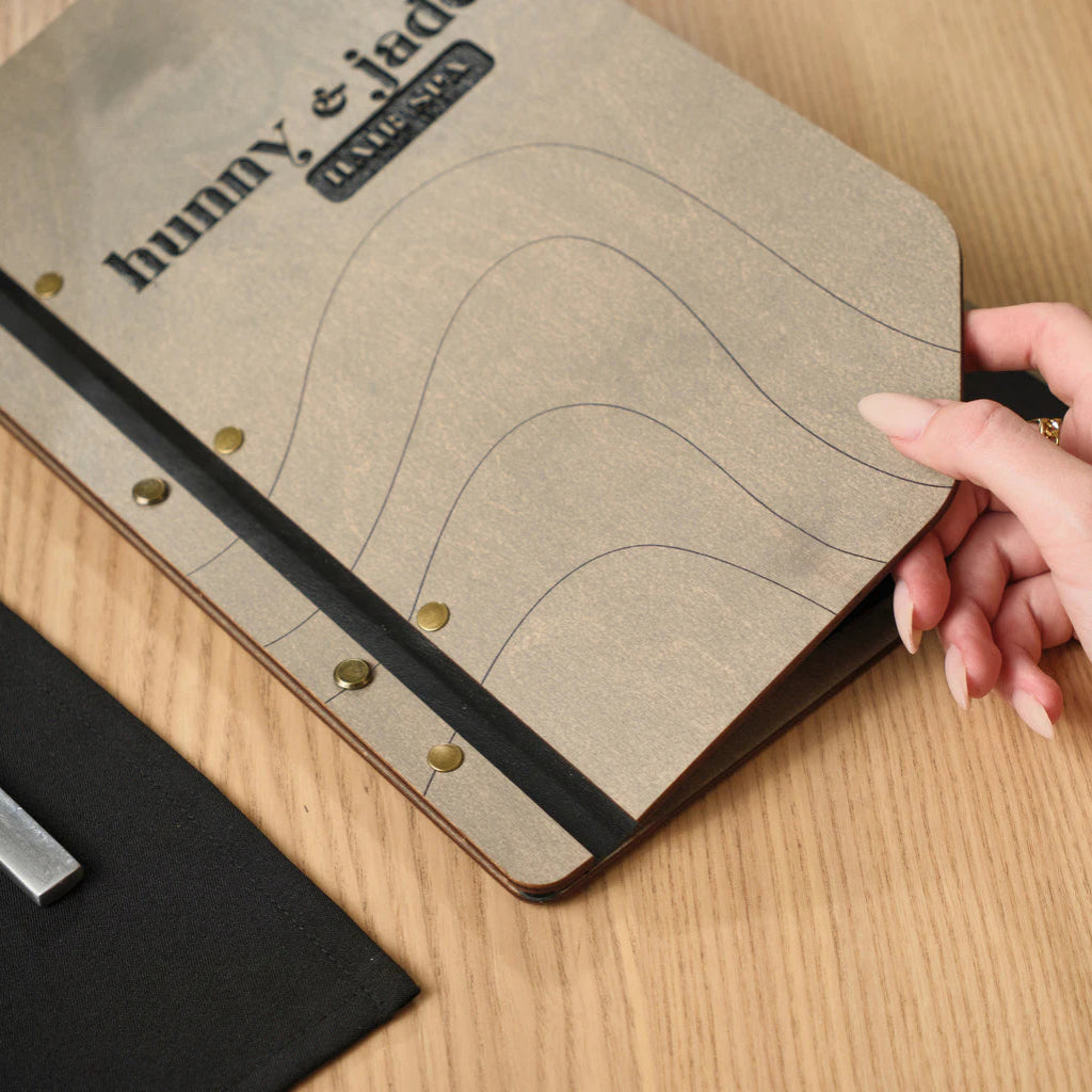 Close-up of a hand opening a custom gray wooden menu board for "Hunny & Jade Hair Spa," showcasing the detailed wavy line engraving and antique brass screw posts