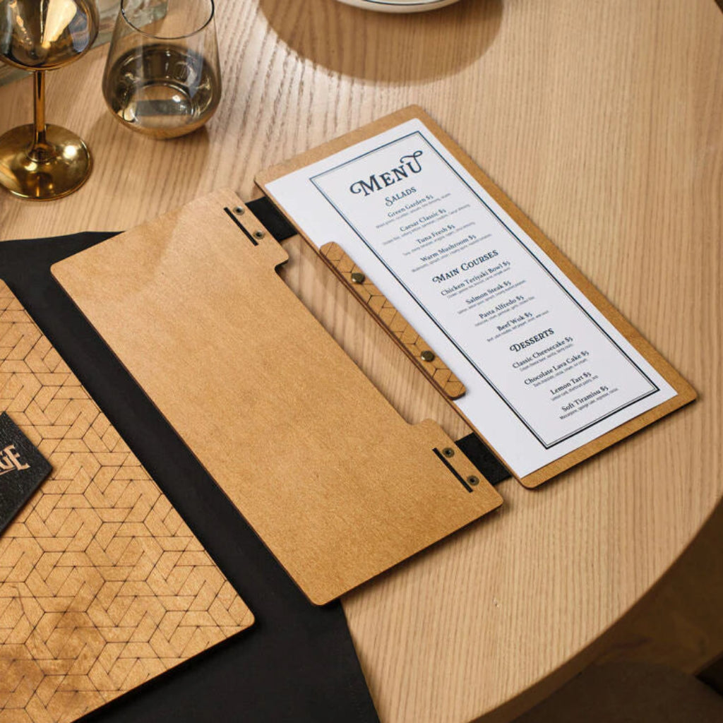 An open oak-colored wooden menu folder lying on a table, displaying a white paper menu with sections for Salads, Main Courses, and Desserts. The internal page is held by a thin wooden strip with brass screws.