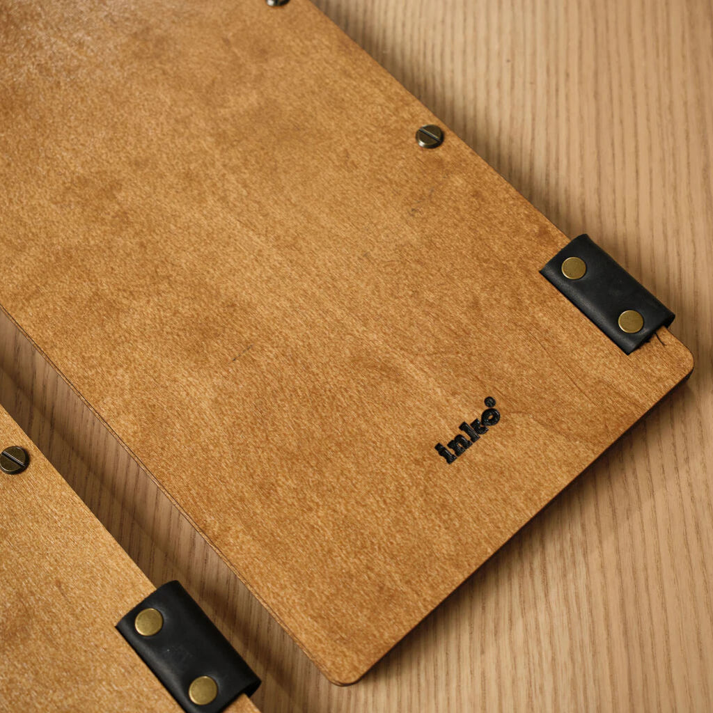 A close-up view of the back of an oak-colored wooden menu folder, showing the natural wood grain and a small black "inko" logo UV-printed at the bottom. Black leather straps with brass rivets and metal binding screws are visible along the edges.