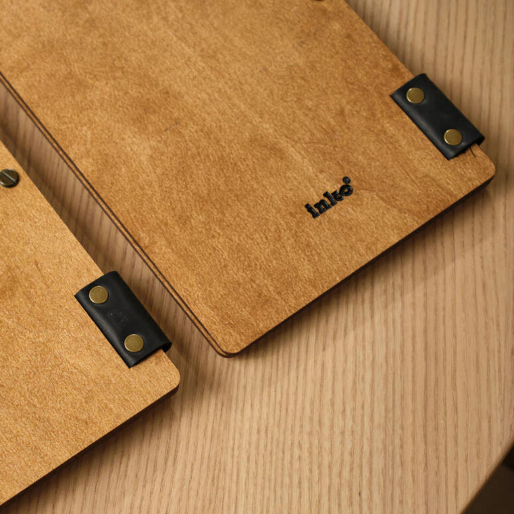 A close-up of the back of the wooden menu folder, showing the smooth oak grain, black leather binding straps with gold rivets, and a small black "inko" logo at the bottom.