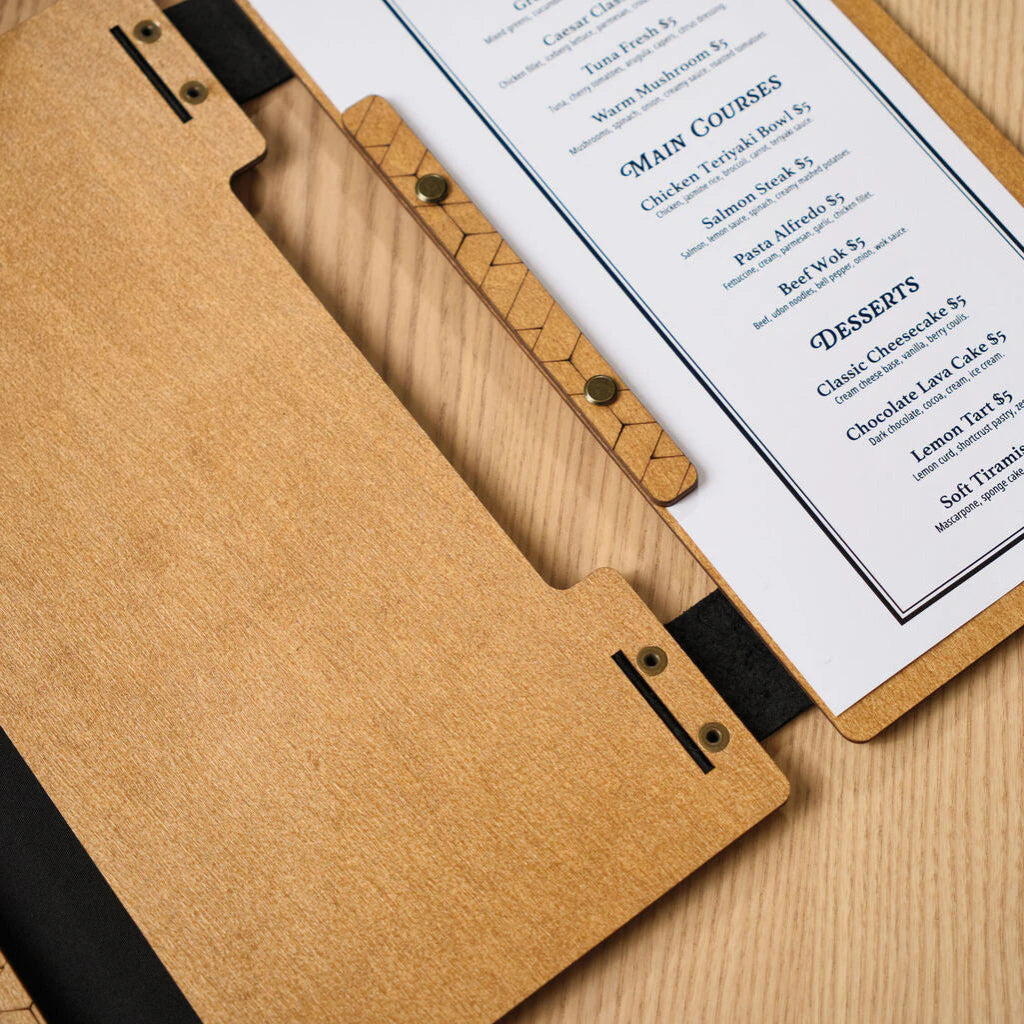 An overhead shot of an open wooden menu folder with a light oak finish, highlighting the black leather hinge and the "Main Courses" and "Desserts" sections of the printed menu.