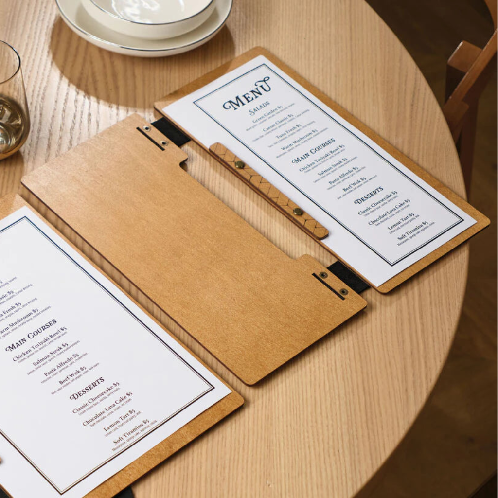 Two open oak wooden menu folders on a light wooden table, showing a clean menu layout. The design features a mix of natural wood textures and modern geometric engravings.