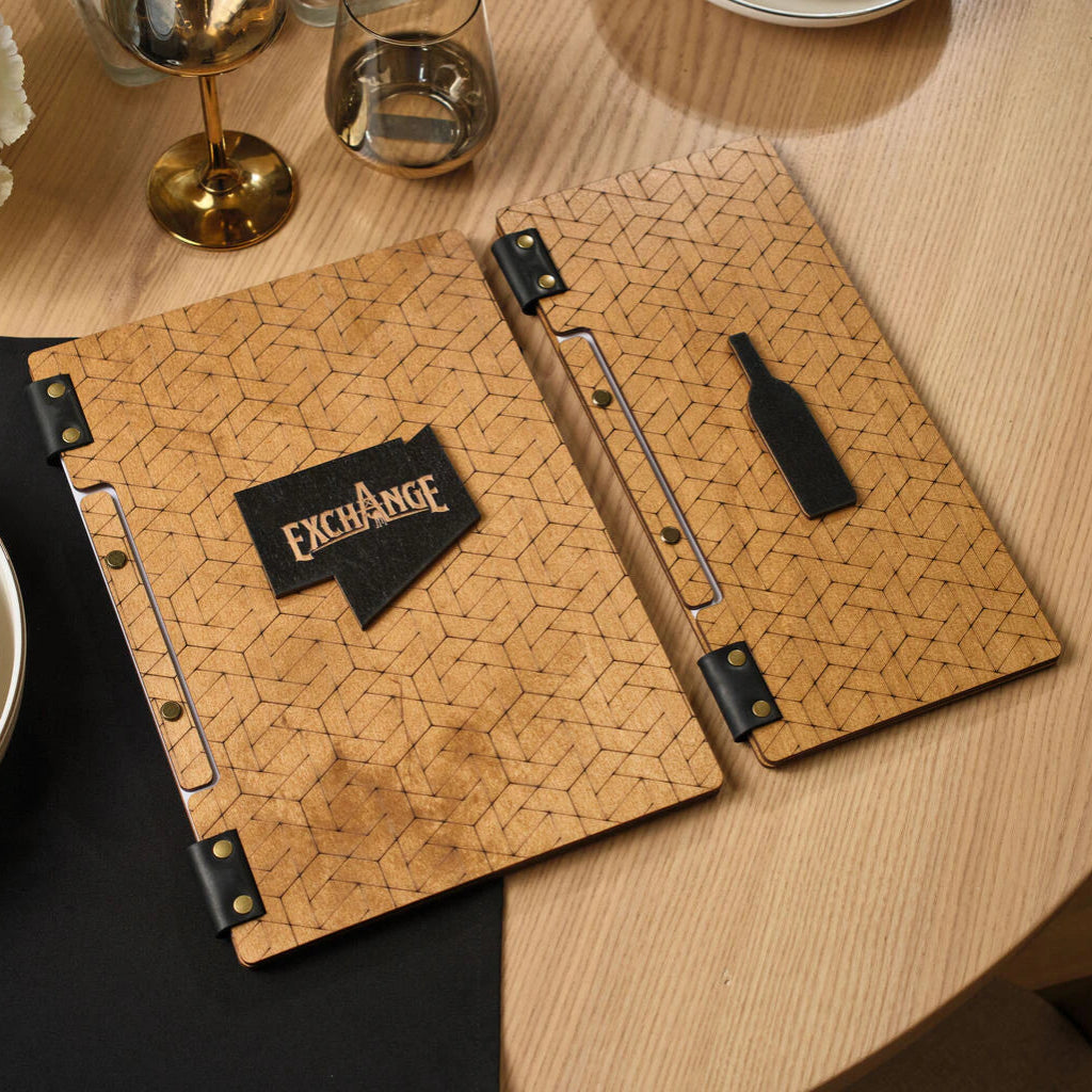 Two wooden menu folders with an oak finish placed side-by-side. One features a black 3D geometric speech bubble with the "EXCHANGE" logo, while the other has a black decorative bottle-shaped inlay.