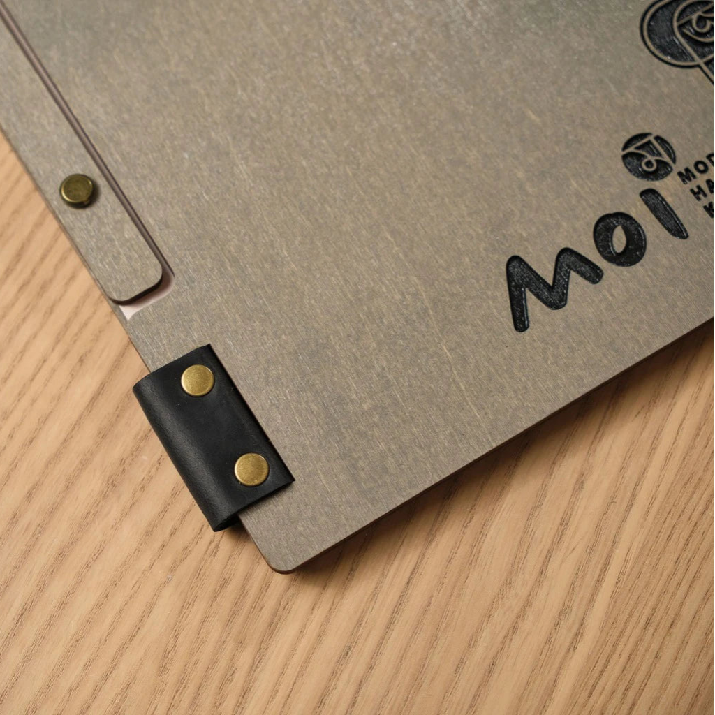 Extreme close-up of a grey wooden menu folder featuring a black leather strap hinge and bronze metal rivets. The shot highlights the premium wood grain texture and custom laser-engraved "Moi" branding for a professional restaurant look