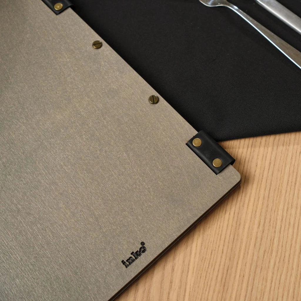 Detailed close-up of the back of a grey wooden menu folder featuring an engraved "inko" logo. This handcrafted wood menu cover highlights the premium textured finish, unique black leather strap accents, and professional bronze bolt binding