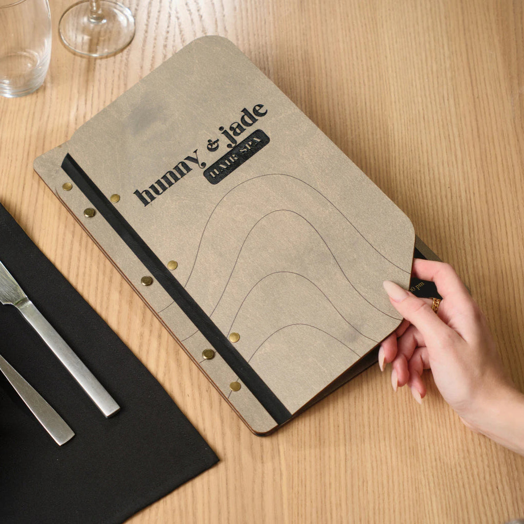 A hand holding the corner of an engraved gray wooden spa menu for "Hunny & Jade Hair Spa," featuring artistic wavy lines and gold-tone fasteners