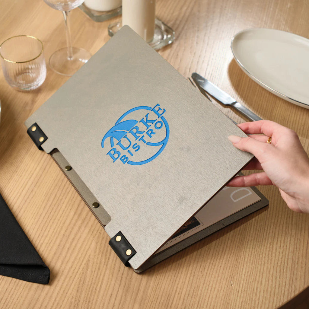 A person's hand lifting the front cover of a grey ash wood menu folder, revealing the printed menu pages inside. The setting is a bright, modern restaurant table.