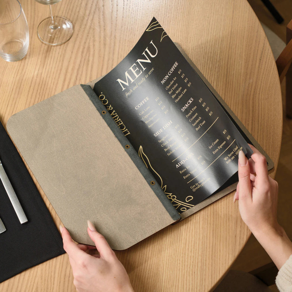 A person holding open a handcrafted gray wood menu folder, displaying a sleek black menu page with gold lettering on a restaurant table.