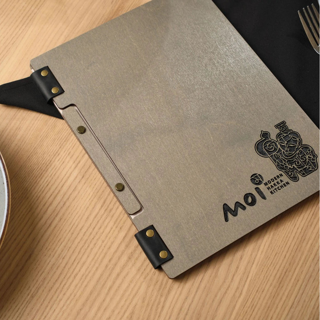 Premium grey wooden menu folder for "Moi Modern Hakka Kitchen" featuring custom laser-engraved branding. This handcrafted wood menu cover includes a unique black leather strap binding with bronze hardware, presented in a professional restaurant setting