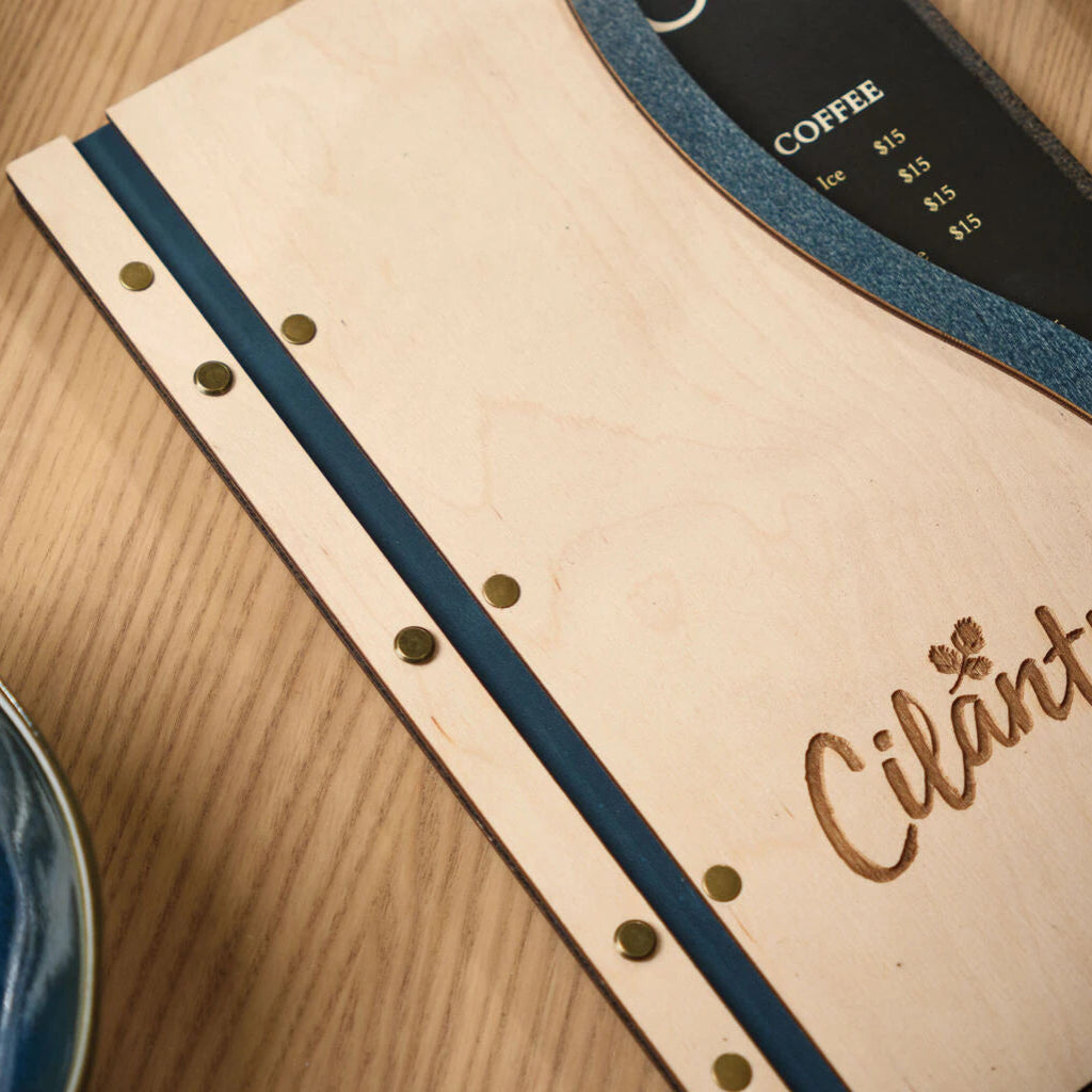 Side-angle shot of a premium wood menu folder, showcasing the stylish contrast between the ivory faceplate and the denim-textured blue spine with gold-tone hardware