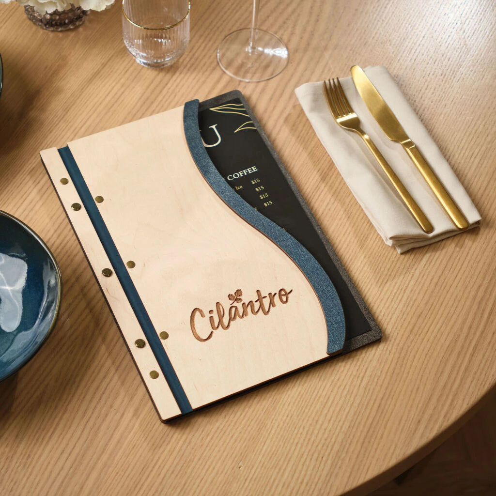 Custom wavy wood menu board in Ivory and Jeans blue, featuring an engraved "Cilantro" logo and brass fasteners, styled on a restaurant table with gold cutlery