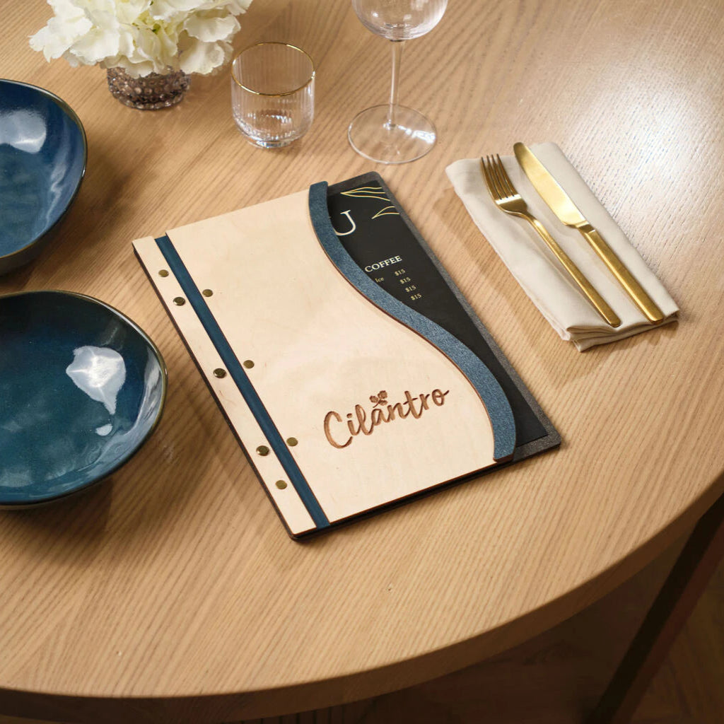 Custom wavy wood menu board in Ivory and Jeans blue, featuring an engraved "Cilantro" logo and brass fasteners, styled on a restaurant table with gold cutlery