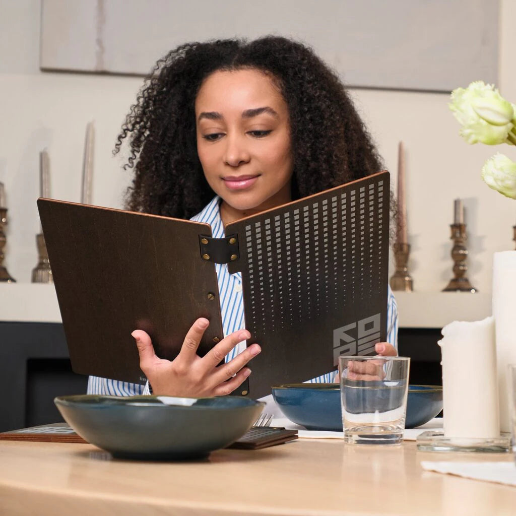 A woman with curly hair is shown from the front, holding open a dark wooden menu folder at a dinner table. The front of the folder features a grey UV-printed geometric square pattern and the word "KORE".
