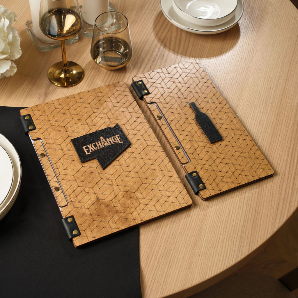 A set of wooden menu and wine list covers featuring an engraved geometric pattern, leather accents, and the "EXCHANGE" logo on a set restaurant table.