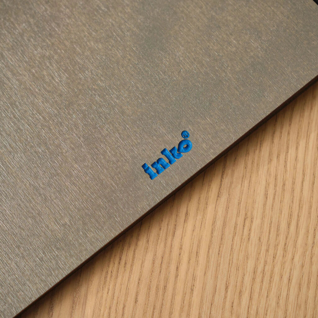 A macro shot of the bottom corner of a grey wooden menu folder, focusing on the debossed blue "inko" logo against the metallic-grey wood grain.