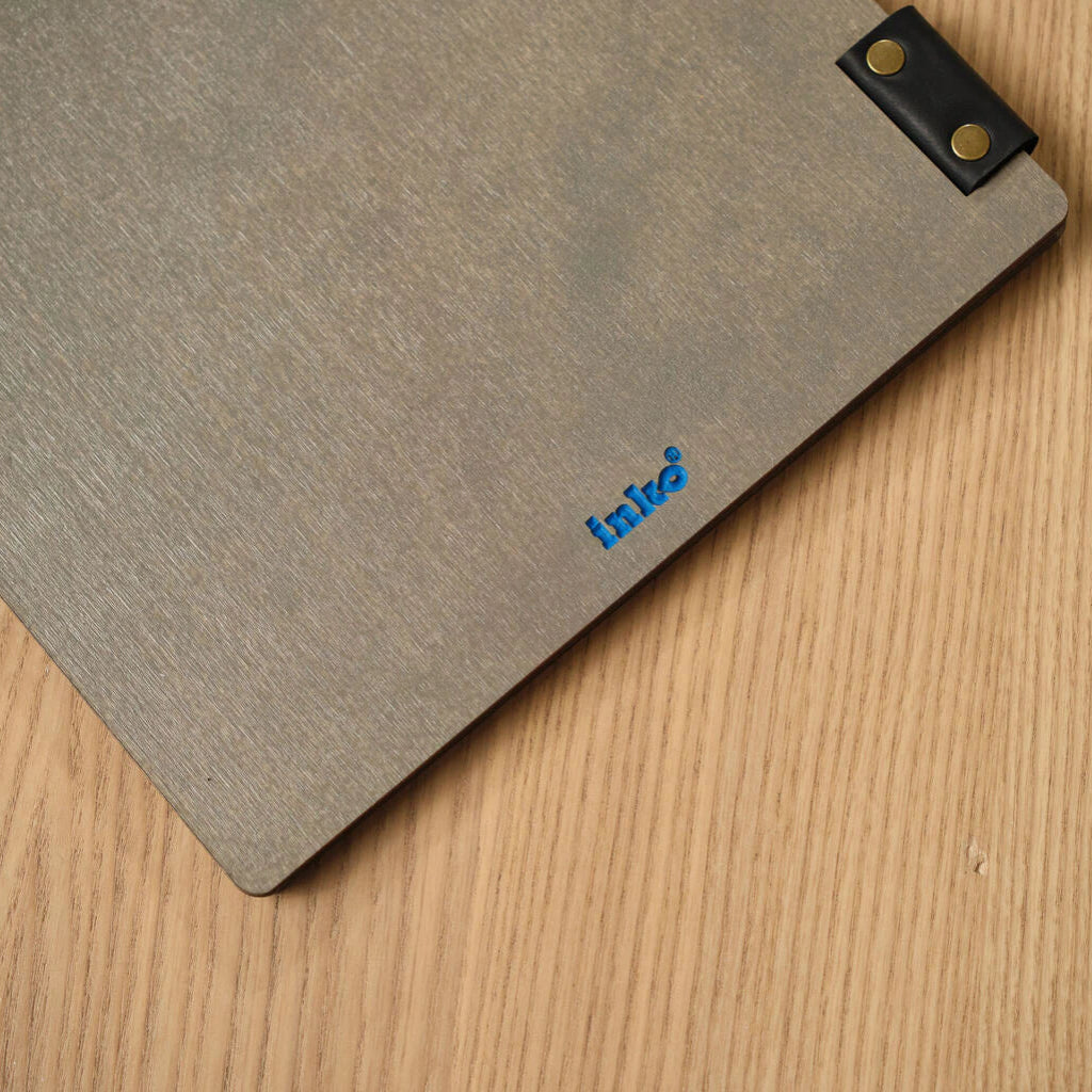 A close-up shot of the back of a grey ash wood menu folder, featuring a small blue debossed "inko" logo in the bottom right corner and a black leather binding strap with brass rivets.