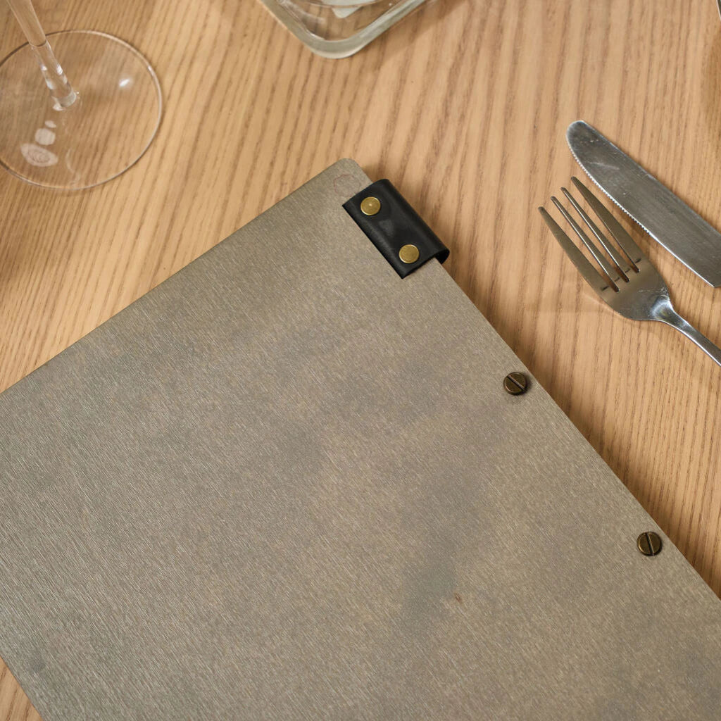 An overhead view of the back of a grey wooden menu folder lying on a light wood table next to a silver fork and knife. The image highlights the natural wood grain and industrial-style brass screw-post binding