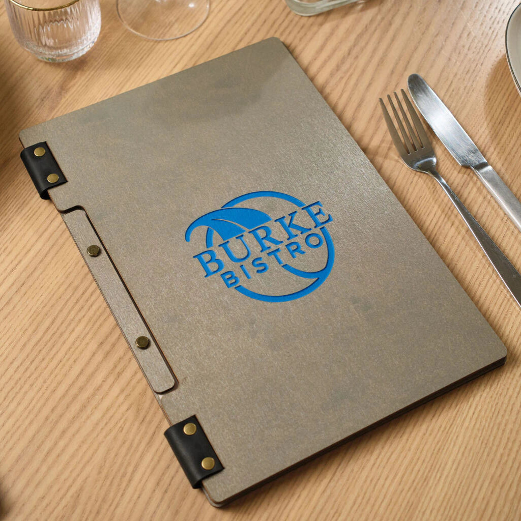 A closed grey ash wood menu folder on a restaurant table featuring a vibrant blue "BURKE BISTRO" circular logo. The folder is bound with black leather loops and gold-toned rivets.