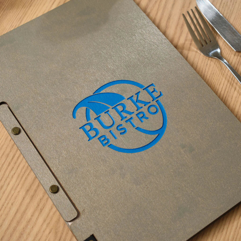 A close-up view of the front cover of a grey wooden menu, showcasing the detailed blue debossed "BURKE BISTRO" branding and the brass hardware on the binding.