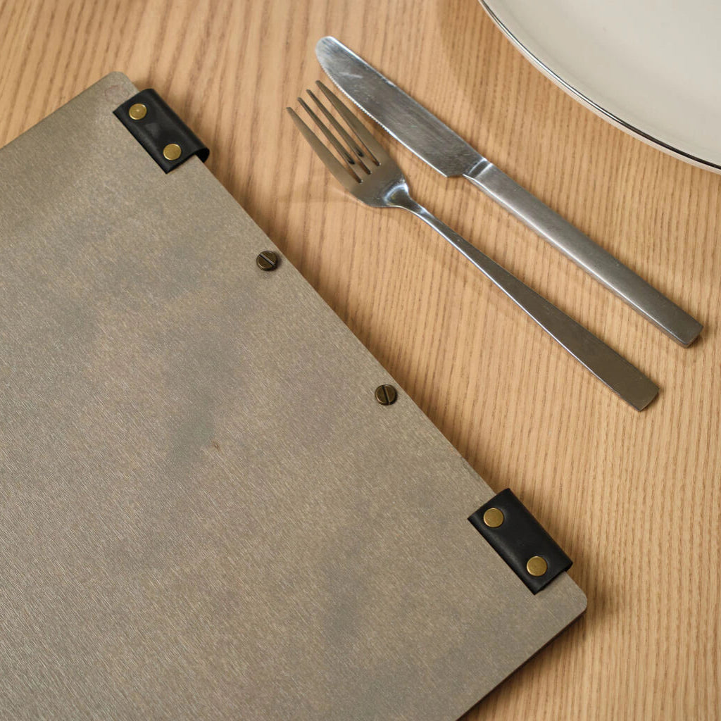 Top-down view of the back of a grey wooden menu folder lying on a light wooden table next to a silver fork and knife, emphasizing a clean and modern restaurant aesthetic.
