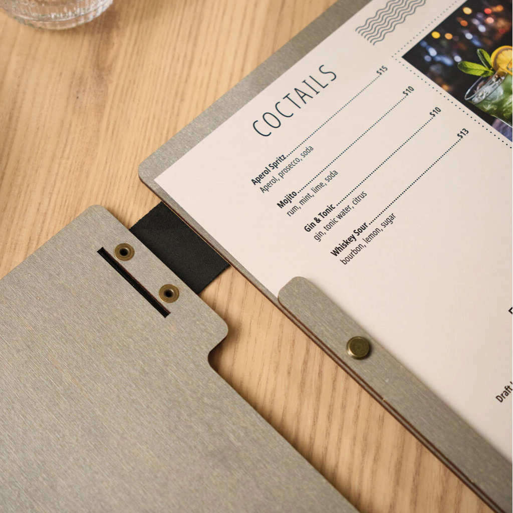 A detailed view of the interior binding of a wooden menu, showing the black leather hinge and the brass screws that hold the cocktail list and menu pages in place.