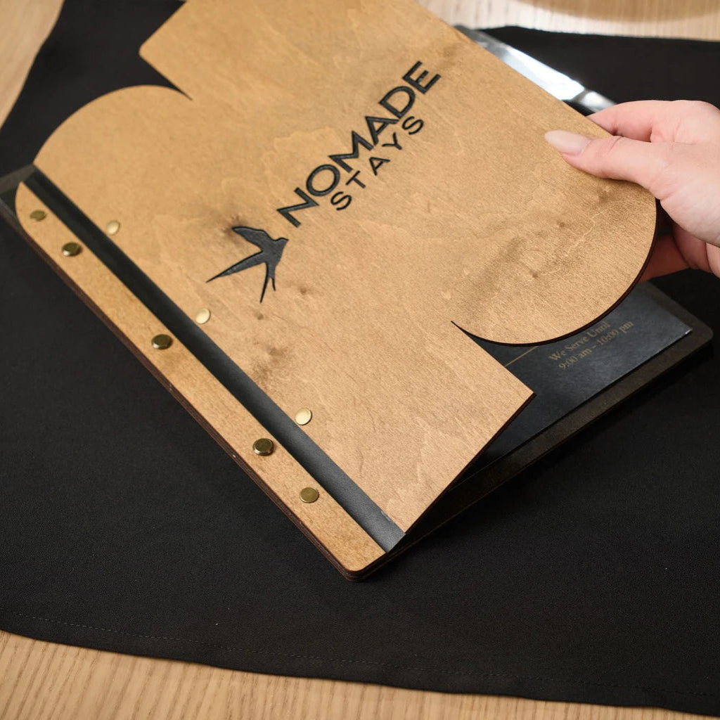 Close-up of a natural oak wooden menu folder with "NOMADE STAYS" engraving, being held by its custom-shaped edge