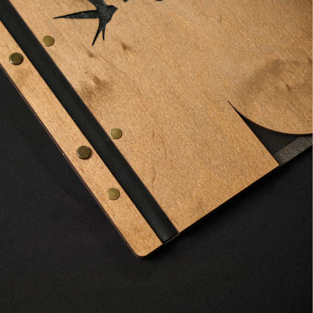 Close-up of a wooden menu folder's binding, showcasing brass screw posts against natural oak and a black spine.