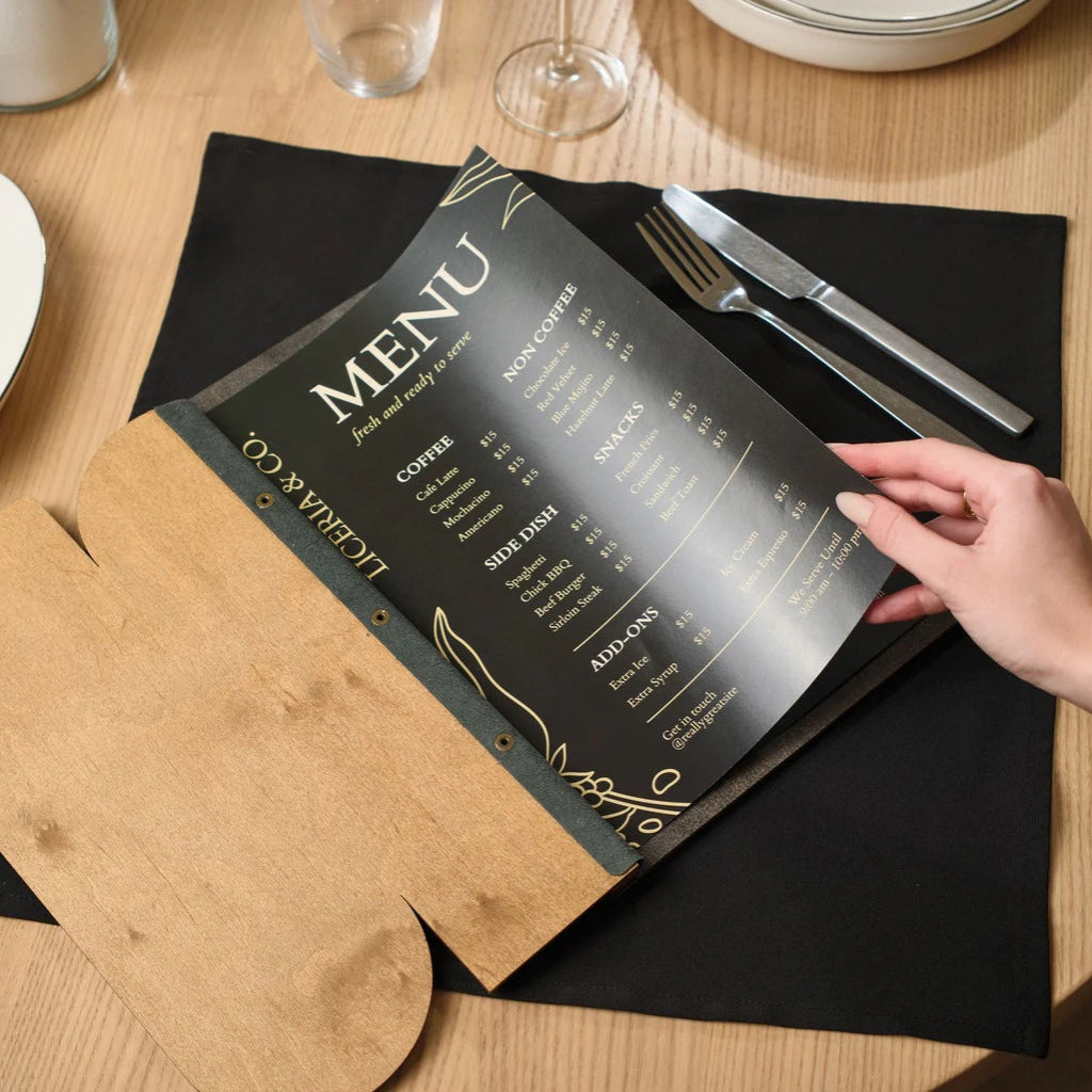 A hand opening a wooden menu folder with a unique custom cutout, crafted from natural oak, showing the internal black menu sheet.