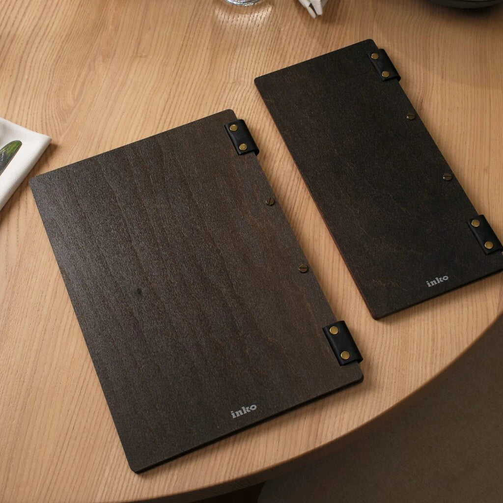 A top-down view of two wenge-colored wooden menu folders lying on a light wood table. The folders feature natural wood grain, black leather hinges with brass rivets, and a small silver "inko" logo at the bottom.