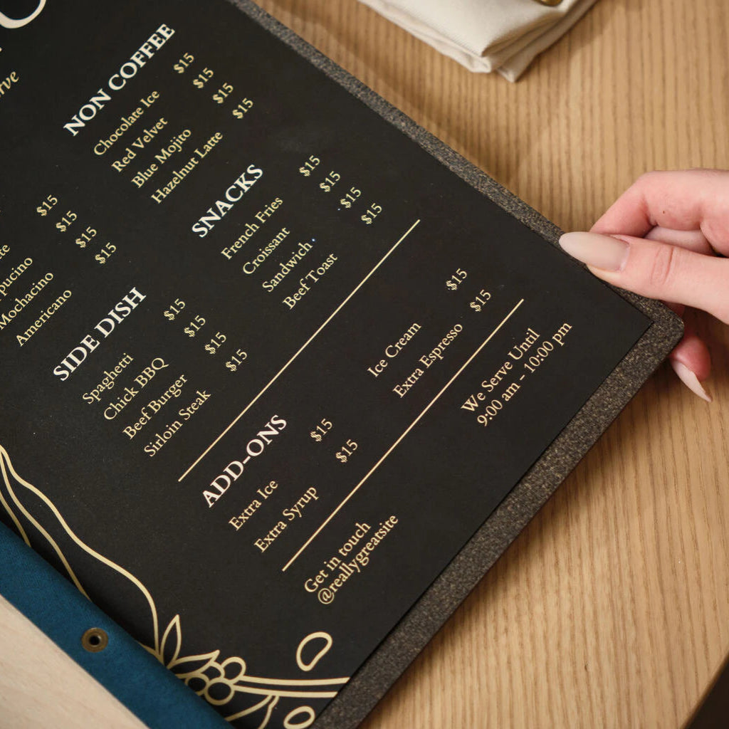 A hand holding the corner of a black restaurant menu insert, showcasing the elegant typography and the dark textured backing of the wooden menu folder