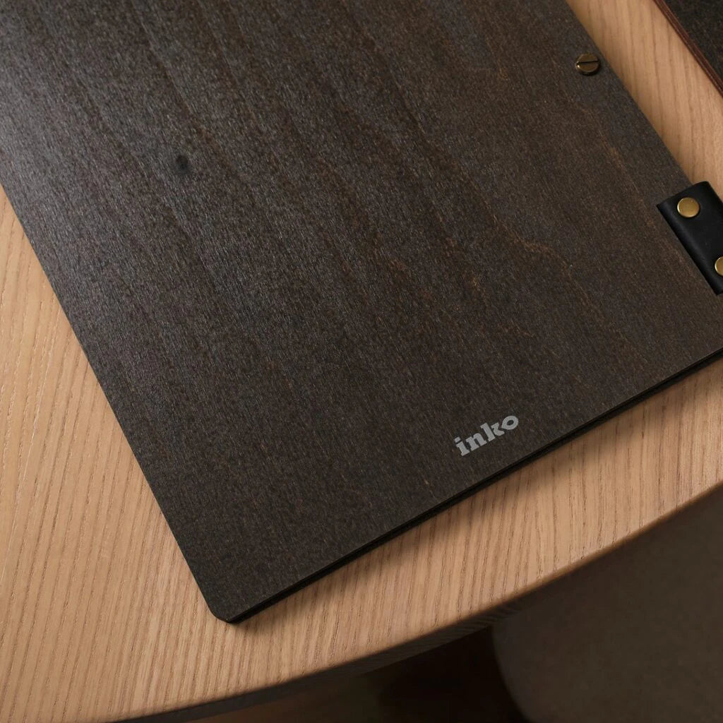 A close-up shot of the corner of a dark wenge wooden menu folder. The image highlights the natural wood texture, a brass binding screw, and the small, silver "inko" logo printed near the bottom edge.