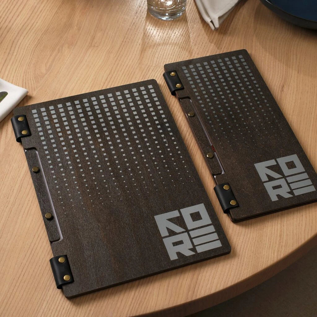 Two wenge-colored wooden menu folders featuring a grey UV-printed gradient square pattern and the "KORE" logo. The folders are held together with black leather hinges and gold-toned brass rivets.