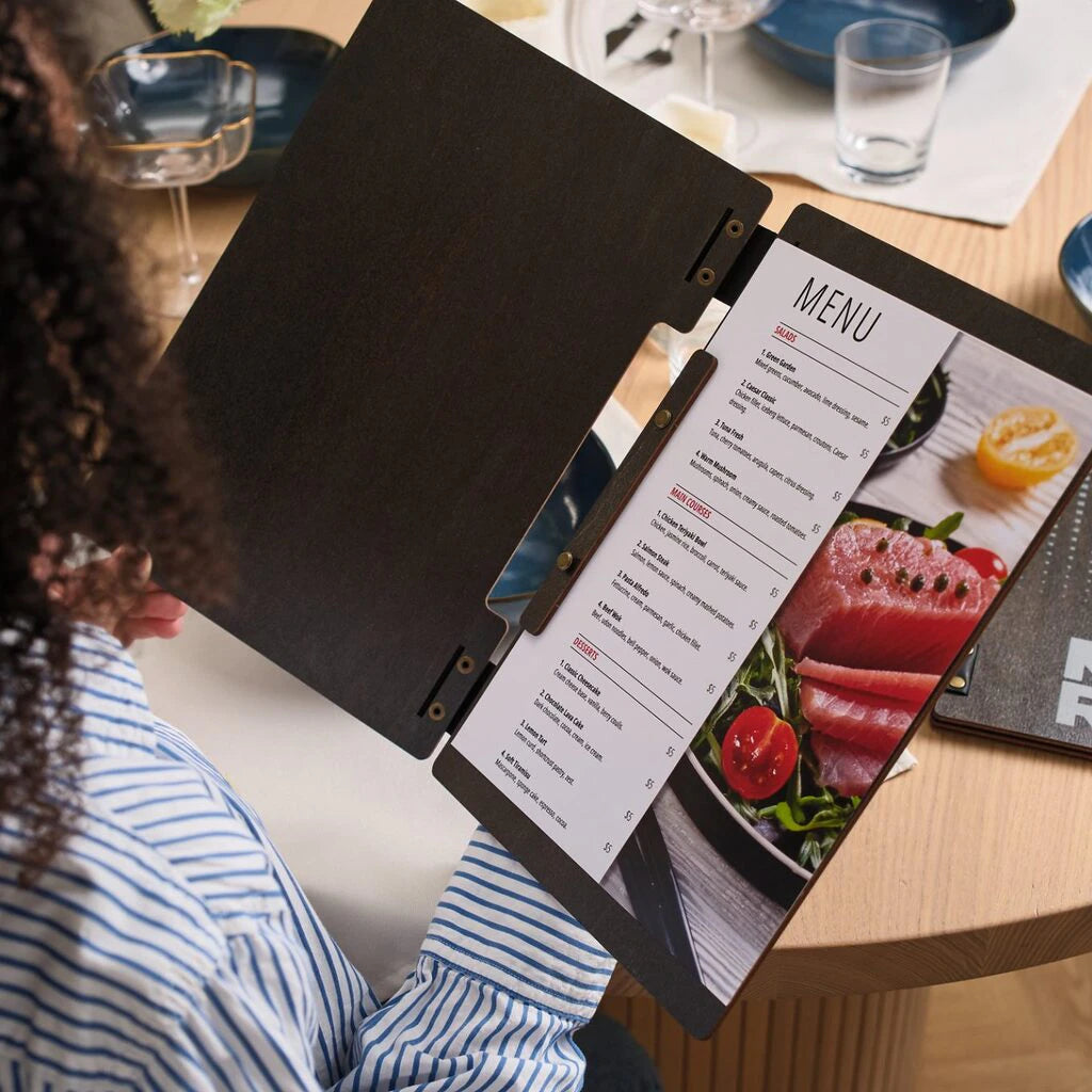A person wearing a blue and white striped shirt holds an open wooden menu folder, looking at a printed list of dishes. The menu features sections for Salads, Main Courses, and Desserts alongside a large image of fresh fish.