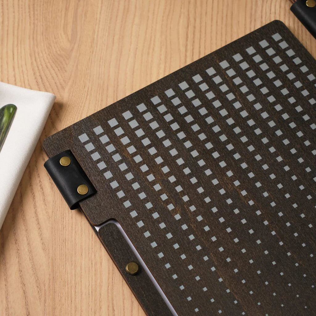 A close-up, top-down view of a wenge-colored wooden menu cover featuring a grey UV-printed pattern of small squares. The cover is accented with a black leather strap secured by gold brass rivets.