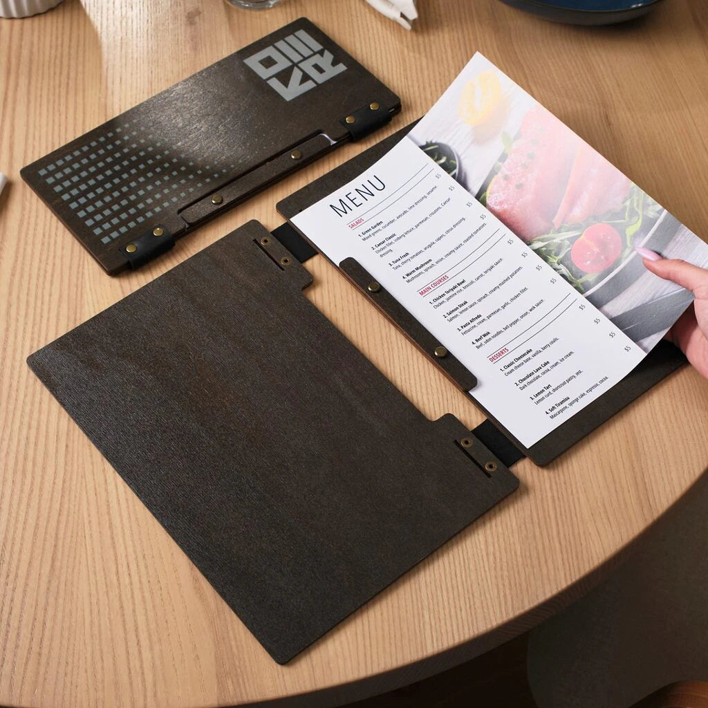 An overhead shot of an open wenge-colored wooden menu folder. A person's hand is visible, turning the page of a printed menu that includes a vibrant photo of a tuna dish.