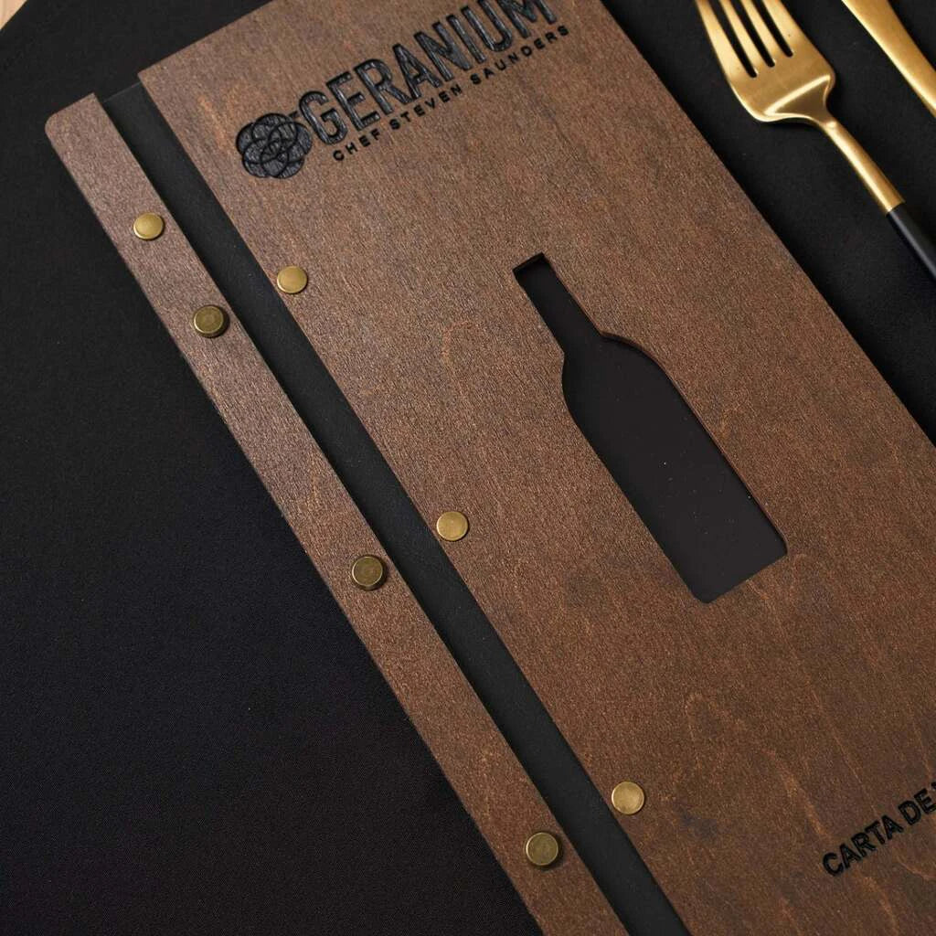 A perspective shot of the front cover lying on a black surface, showcasing the decorative wine bottle cutout and the "GERANIUM CHEF STEVEN SAUNDERS" logo engraved at the top.