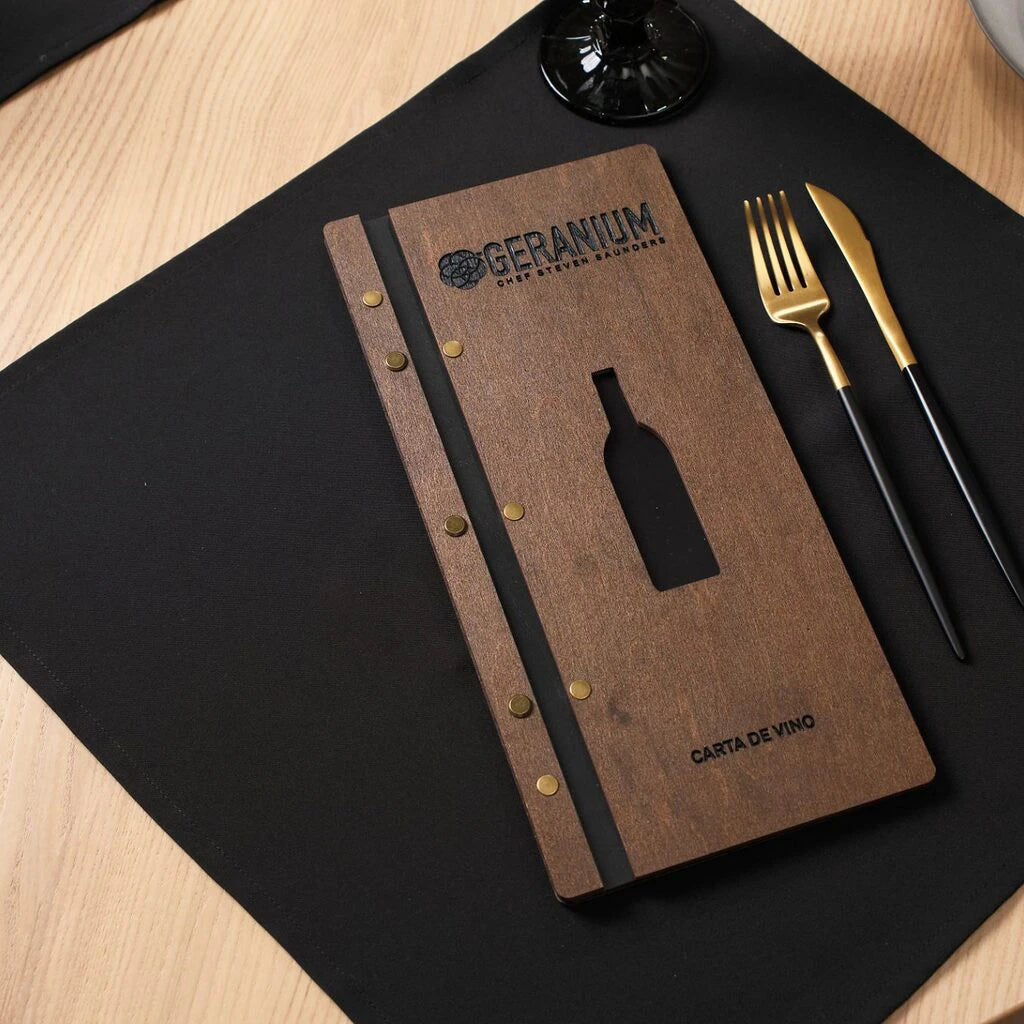 A walnut-colored wooden menu folder lying flat on a black napkin, featuring a central cutout in the shape of a wine bottle and the engraved text "GERANIUM" and "CARTA DE VINO".