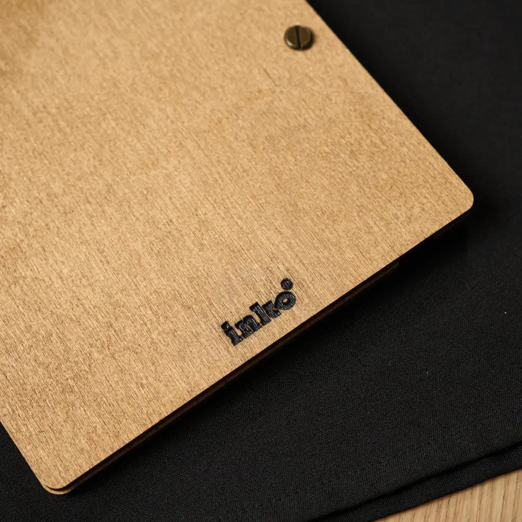 A close-up of the bottom corner of the oak menu's back cover, showing the black "inko" brand logo engraved into the wood grain next to a dark brass binding screw.
