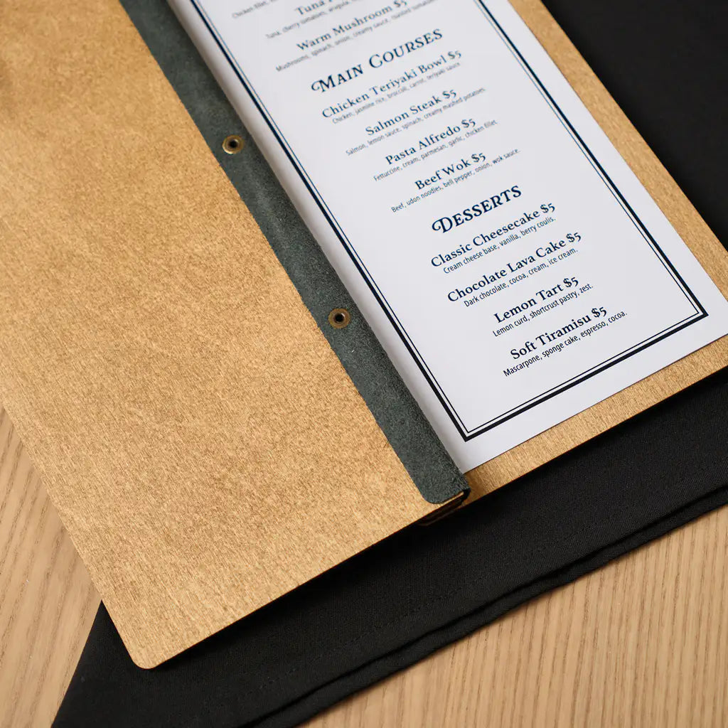 A high-angle close-up focusing on the "Desserts" section of the menu and the dark gray spine strip with brass rivets that keeps the pages perfectly aligned within the wooden cover.binding.