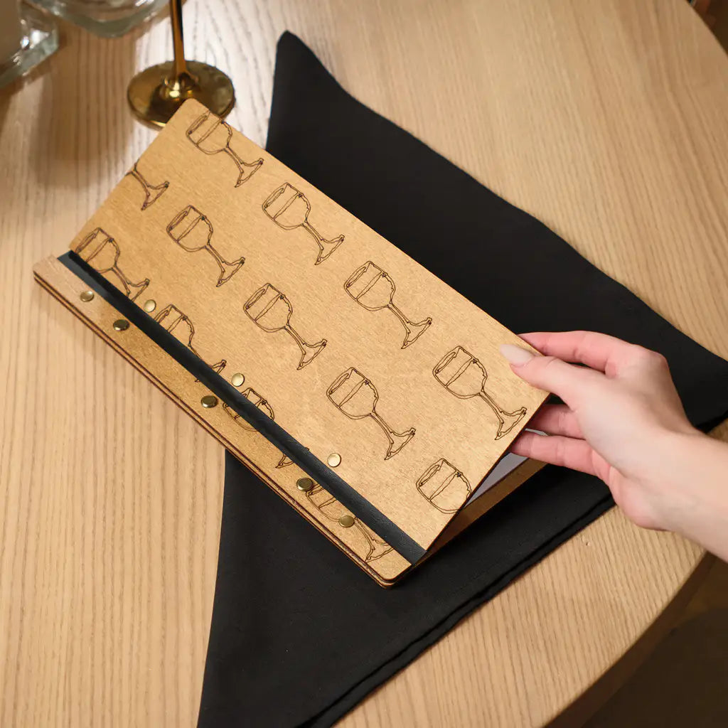 : A person’s hand lifting the front cover of the menu folder, which features a decorative engraved pattern of wine glasses.