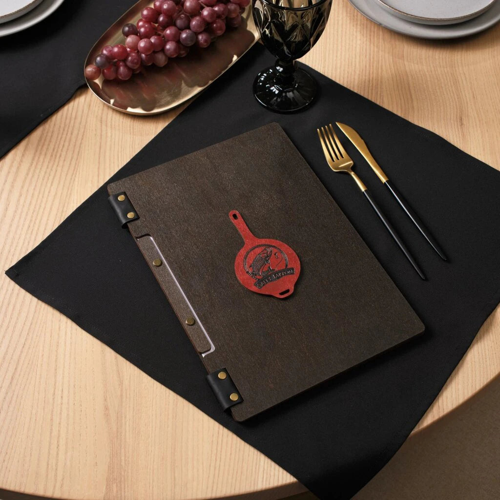 A closed wenge-colored wooden folder on a black napkin, decorated with a red leather frying-pan-shaped logo and brass details