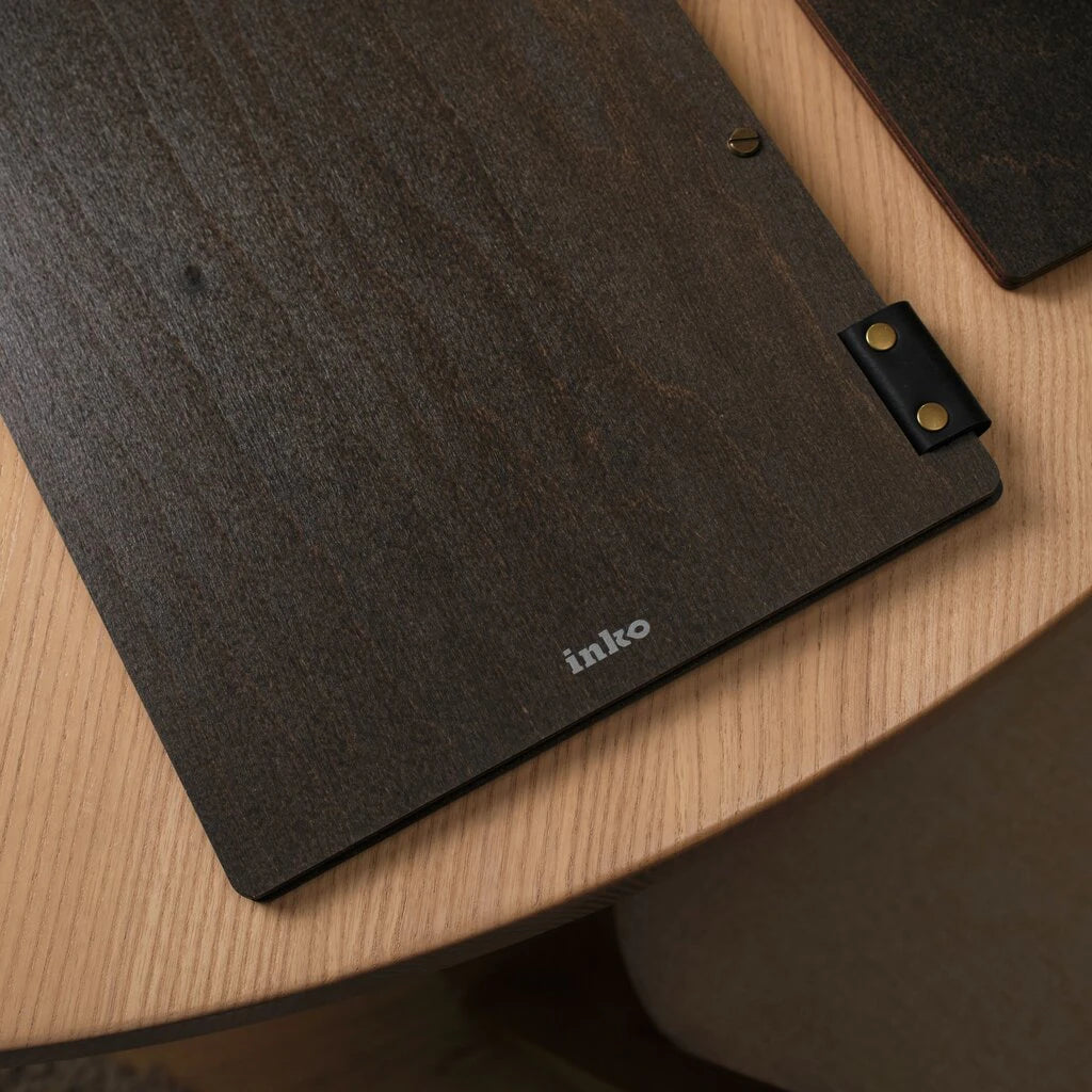 Dark wood menu holder with leather hinges, brass rivets, and a white "inko" logo, shown in a close-up detail shot
