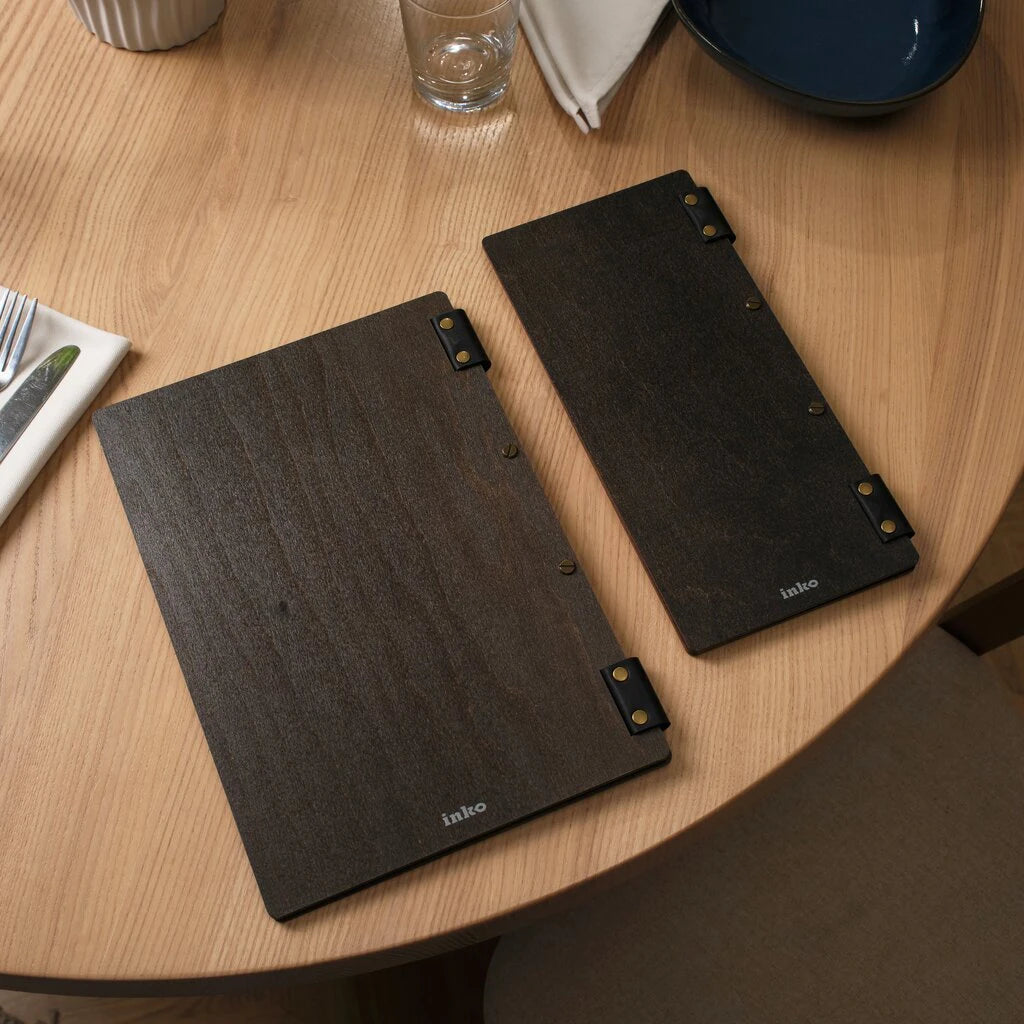 Two dark wood menu holders in different sizes: a standard portrait folder and a slim elongated version. Both feature black leather hinges with brass rivets and a screw-post binding system. The minimal design includes a subtle white "inko" logo on the bottom of each dark-stained wooden cover