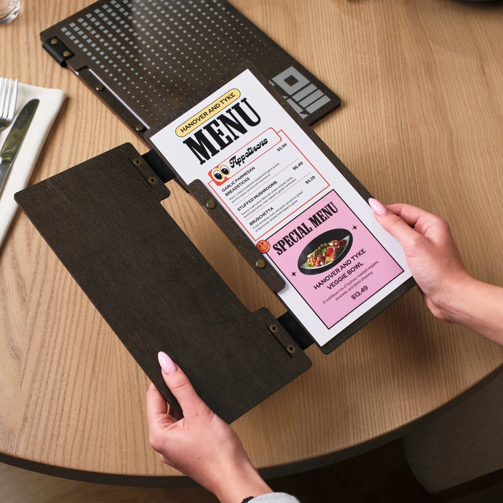 A slim-format dark wood menu holder held open by a person to display a "Special Menu" sheet. The folder is constructed with two wooden panels connected by flexible black leather hinges and brass rivets. The internal pages are secured with a visible bolt-binding system on the right-hand panel