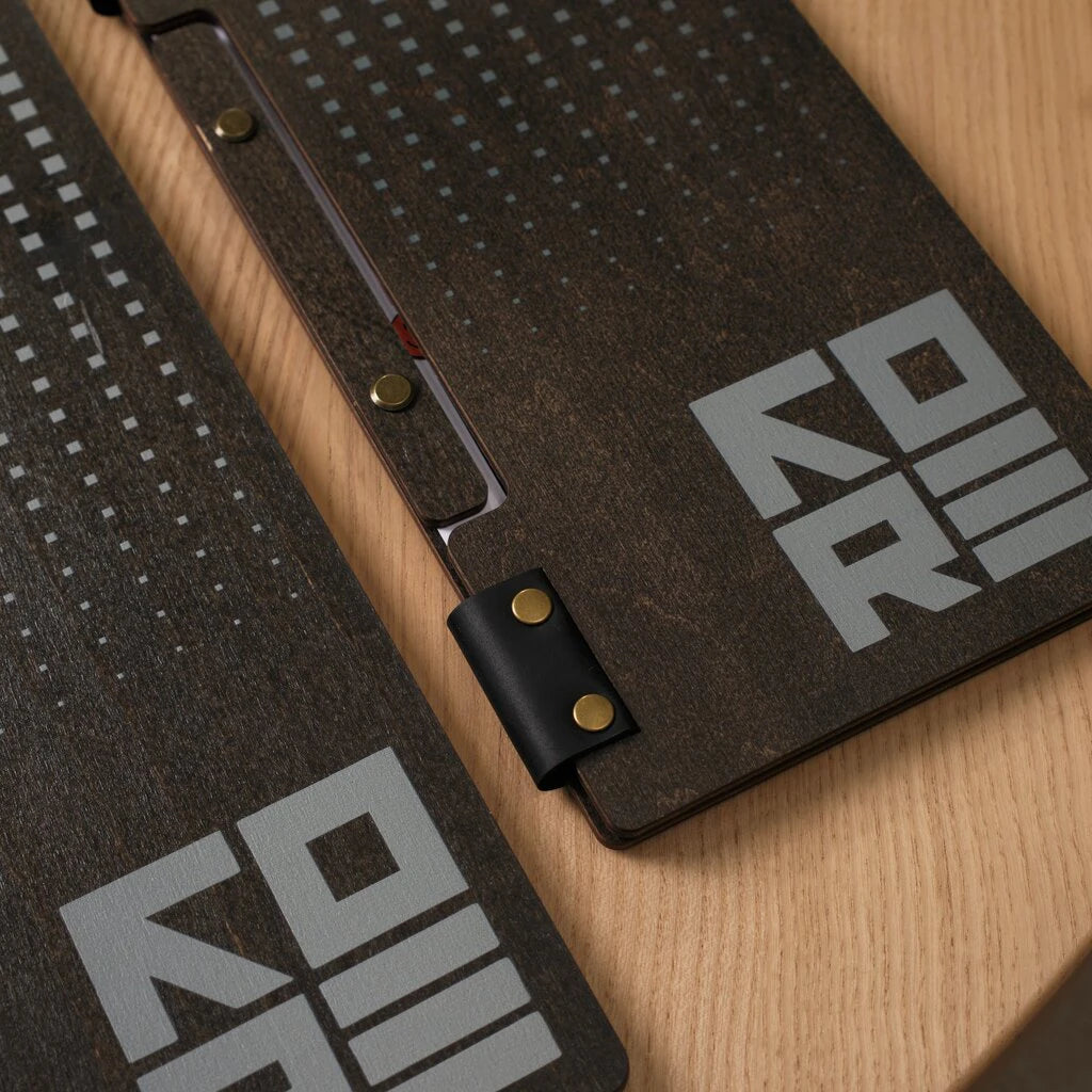 Close-up of two dark wooden menu covers on a light oak table. The image highlights the textured wood grain, a laser-engraved grey "KORE" logo, and a geometric square pattern. The folders are held together by black leather straps with gold-tone brass rivets and a screw-post binding system