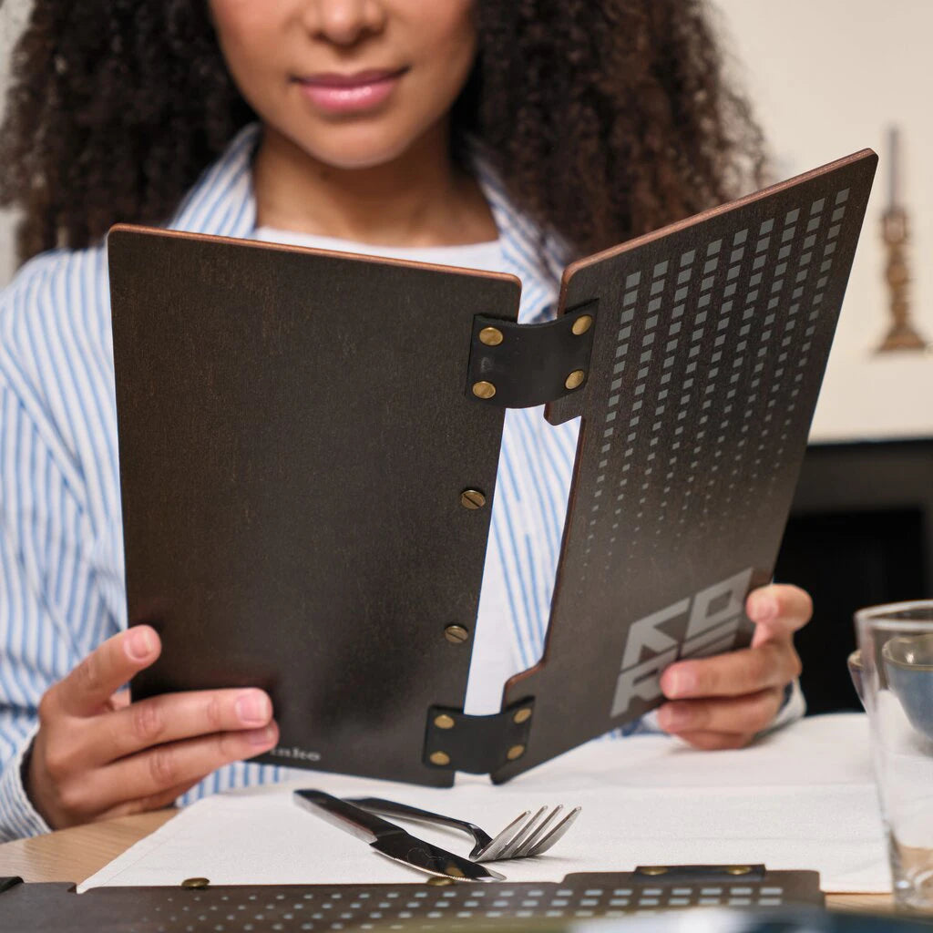 A dark-stained wooden menu cover held by a customer. The folder features a unique split-panel design connected by two black leather hinges with brass rivets. The wooden surface includes a laser-engraved geometric pattern and branding, with a vertical bolt-binding system for the internal pages