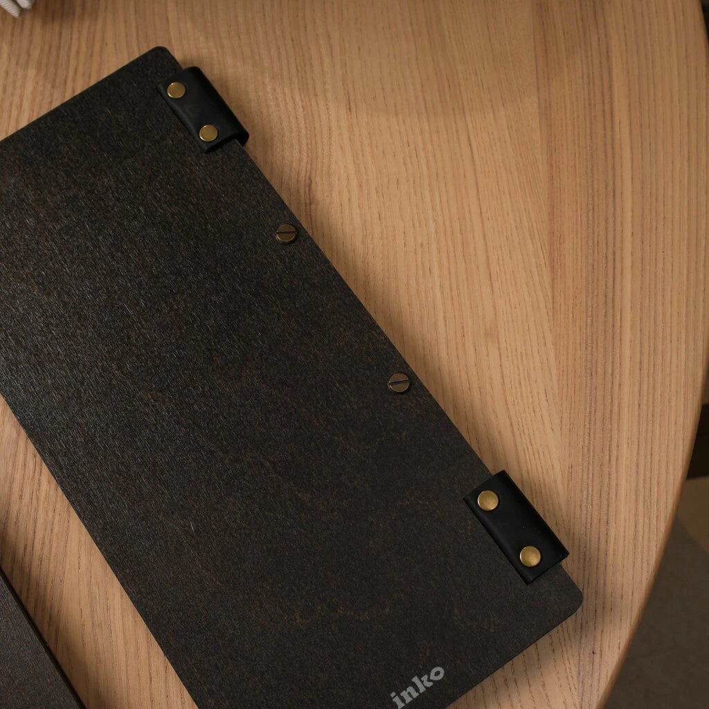 Close-up of a slim dark wood menu holder with a tactile grain finish. The design features a flexible spine with black leather hinges and gold-tone brass rivets. Two central screw-post bolts allow for secure paper insertion, and the "inko" brand logo is subtly printed at the bottom