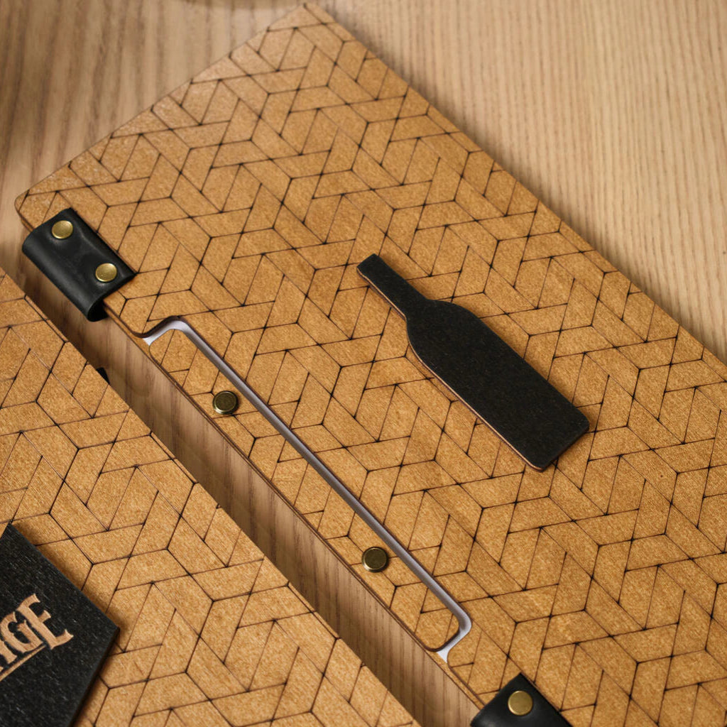 A detailed top-down shot of a wooden menu folder featuring an all-over laser-engraved geometric pattern of interlocking shapes. A black decorative inlay in the shape of a wine bottle is centered on the cover, which is bound with black leather and brass hardware.