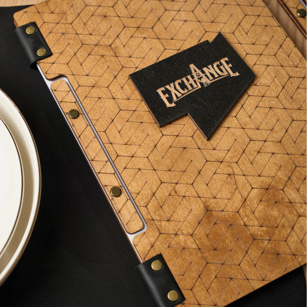 Side view of a patterned wooden menu folder, showing the raised "EXCHANGE" logo and the internal sheet-holding system