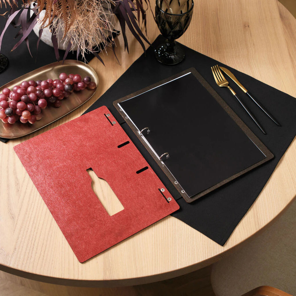 Top-down view of an open chili-red wooden menu folder on a restaurant table, featuring black pages, gold cutlery, and a bowl of grapes